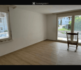 Empty room with wooden flooring, white walls, large window with blinds, and a single wooden chair near the sliding glass door leading to a balcony.