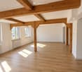 Bright, empty room with wooden flooring and exposed wooden beams on the ceiling. Large windows allow natural light to fill the space, and there are white walls and a radiator visible.