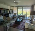 Elegant living room with large windows offering a scenic view, featuring a cream-colored sofa, glass coffee table, glass dining table with chairs, and a patterned area rug.