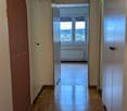 A long, narrow hallway with wooden flooring and white walls, leading to a room with large windows and a radiator.