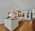 Bright kitchen with white cabinets, wooden countertop, and hardwood flooring. Features a sink, faucet, table, fan, oven, bookshelf, and clock.
