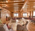 Cozy living room with wooden beams, exposed wooden ceiling, large windows, floral-patterned sofa and armchair, dining table with chairs, television, and floor lamp.