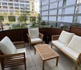 Balcony with wooden furniture including a sofa, chairs, and a small table, surrounded by modern buildings and greenery.