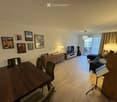 A modern living room with a wooden floor, a sofa, a dining table with chairs, a TV, and several lamps. There are picture frames on the wall and a large window with curtains leading to a balcony.
