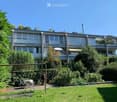 Modern multi-story building with large windows and balconies, surrounded by lush greenery and a well-maintained garden.