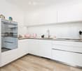 Modern kitchen with white cabinets, stainless steel appliances, and wooden flooring. Features an oven, sink, and various small appliances on the counter.