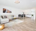 Modern open-plan living and dining area with white walls, wooden flooring, a white sectional sofa, a glass coffee table, a dining table with chairs, and a small kitchen area with white cabinets and a sink.
