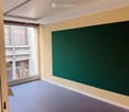 Empty room with light yellow walls, dark grey carpet, large window with view of building exterior, and green chalkboard on one wall.