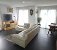 Modern living room with a white leather sofa, coffee table, TV, dining area with chairs and table, potted plants, and decorative items.