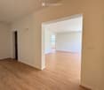 Empty room with light wooden flooring and white walls, featuring an open doorway leading to another room.