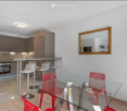 Modern open-plan kitchen and dining area with grey cabinets, a glass dining table surrounded by red chairs, and a bar area with white stools.