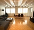 Bright living room with wooden flooring, exposed wooden ceiling beams, large windows with white curtains, a grey sectional sofa, a wicker coffee table, a dining table with chairs, a flat-screen TV on a stand, and a small Christmas tree in the corner.