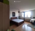 Bright bedroom with wooden flooring, two beds, nightstand, lamp, picture frame, plant, radiator, and a rug.
