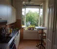 Modern kitchen with white cabinets, stainless steel appliances, and a small dining area with a table and stools. The room features terracotta tiles and large windows with a view of greenery.