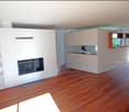 Modern living room with a built-in fireplace, wooden flooring, white walls, and an open kitchen area with a counter and shelves.