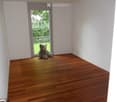 Empty room with wooden flooring, white walls, and a large window with a view of greenery outside.