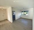 Empty room with white walls, wooden floor, and a door leading to a kitchen. A window with blinds is on the right side.