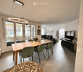 Bright, open-plan living and dining area with large glass doors leading to a balcony. Features a wooden dining table with green chairs, a black leather sofa, a TV stand, and decorative items including a flower vase and picture frames.