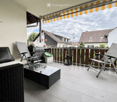 Balcony with a black leather sofa, wicker coffee table, two folding chairs, and a barbecue. The balcony has a wooden railing and a striped awning.