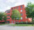 Modern red apartment building with multiple floors, large windows, and balconies. Surrounded by greenery and a paved area.