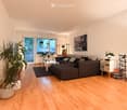 Modern living room with light wooden flooring, large window, grey sectional sofa, coffee table, side table, floor lamp, picture frame, potted plants, sideboard, and shoe rack.