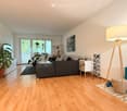 Modern living room with light wooden flooring, grey sectional sofa, coffee table, floor lamp, side table, potted plants, and a picture frame on the wall.