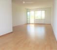 Empty room with light wooden flooring and white walls, featuring large windows and a sliding glass door leading to a balcony.