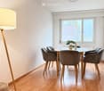 Bright dining area with a round glass table, six grey upholstered chairs, a tall floor lamp, and a small potted plant on the table.