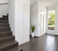 Modern hallway with dark tiled stairs, white walls, and a large window showing greenery outside. A white vase with tall plants is placed near the stairs.