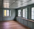 Empty room with wooden flooring, grey walls, and multiple windows with open shutters. The ceiling features exposed beams and skylights.