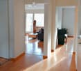 A well-lit hallway with wooden flooring, leading to a living area with a sofa, coffee table, and a view of the outside through large windows.