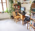 A well-lit study or home office with a wooden desk, computer setup, bookshelf filled with books, and several potted plants near the window.