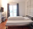 Modern bedroom with a large window and balcony access, featuring a bed, nightstand, lamp, and a TV on a stand.