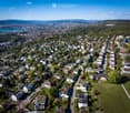 Aerial view of a suburban neighborhood with numerous houses, green spaces, and a distant cityscape with a lake and mountains in the background.