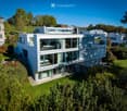Modern two-story house with large glass windows, a raised ground floor, and a well-maintained garden.