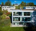 Modern two-story house with large glass windows, a rooftop terrace, and surrounding greenery.