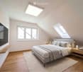 Modern attic bedroom with wooden floors, large windows, skylights, a grey upholstered bed, nightstands, table lamps, and a wall-mounted screen.