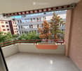 Balcony with concrete floor, red metal railing, and a planter box, overlooking a cityscape with buildings and trees.