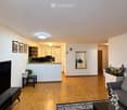 Modern living room with hardwood flooring, a grey sofa, coffee table, TV stand, and a small kitchen area with white cabinets and a countertop. A large mirror is mounted on the wall, and there are framed pictures and plants adding to the decor.