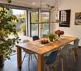 Modern dining room with a wooden table, six chairs, a bowl of fruit, a vase with flowers, and a potted plant. Large windows provide natural light and a view of the garden.