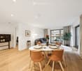 Modern, well-lit living and dining area with wooden flooring, white walls, and large windows. Features a sofa, dining table with chairs, a small cabinet, a table lamp, and decorative plants.