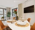 Dining area with a white table set for six, surrounded by orange chairs, a bottle of champagne, and decorative vases with flowers. Large windows offer a view of the city.