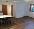 Bright, unfurnished room with wooden flooring, white walls, and a small kitchen area featuring a sink, faucet, and cabinets. A table and chair are positioned near the kitchen area.
