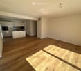Empty room with wooden flooring and white walls, featuring an open kitchen area with cabinets and appliances visible in the background.