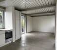 An unfurnished room with a concrete floor, white walls, and a modern kitchen area featuring a built-in oven and countertop. The room has large windows and a high, exposed wooden ceiling structure.