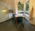 Bright kitchen with white cabinets, stainless steel oven, microwave, and a dining table with chairs, including a baby chair. Large windows provide natural light.