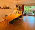 Dining room with a long wooden table surrounded by green chairs, a hanging pendant light, and a shelf with decorative items.