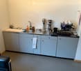 A small, modern kitchen area with a sink, coffee machine, coffee grinder, and various kitchen items on the counter. The cabinets are light blue, and there is a black leather chair in the foreground.