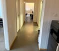 A long, narrow hallway with light-colored walls and a smooth, light-colored floor. The hallway has several doors on both sides, leading to different rooms. There is a printer on a stand to the right, and a glimpse of an office area at the end of the hallw