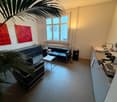Modern living room with a black leather sofa, a coffee table, a cabinet, a coffee machine, and a sink area. The room features large windows, a radiator, and a plant in the corner.